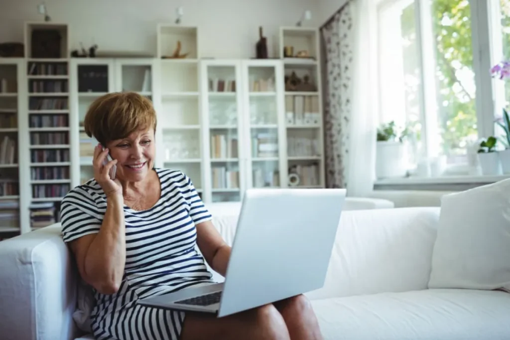 Smiling older woman in a striped dress sits on a white sofa, talking on her phone while looking at a laptop in a sun‑lit living room.