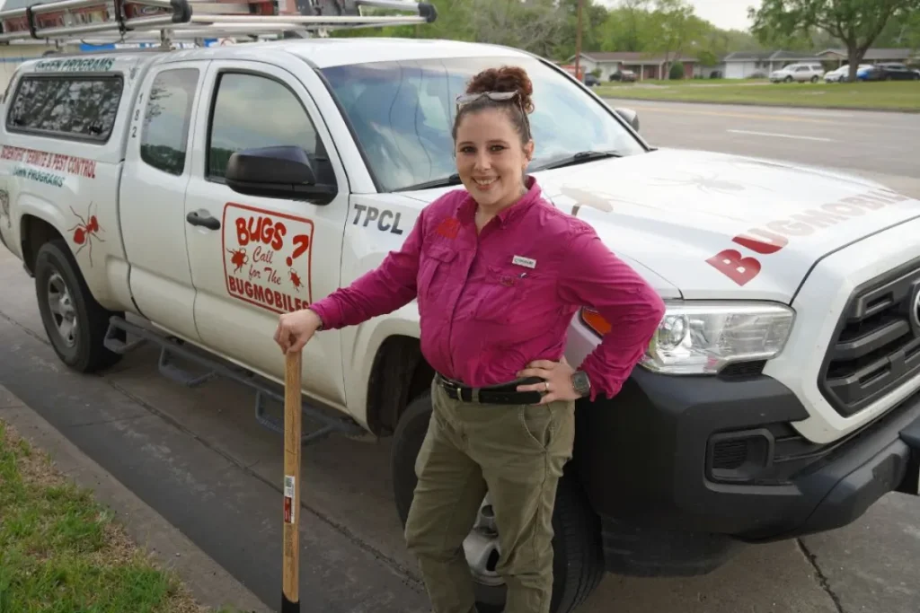 Female pest‑control technician in a bright pink shirt leans on a “Bugmobile” pickup, holding a wooden‑handled tool on a suburban street.