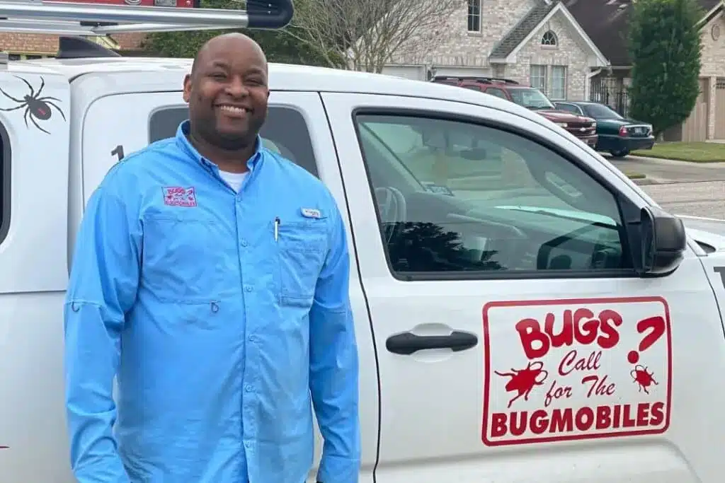 Smiling pest control worker standing beside a white Bugmobiles service truck in a residential neighborhood.