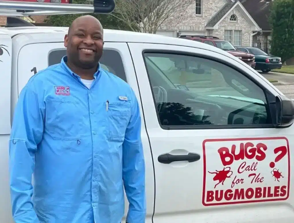 Smiling pest control worker standing beside a white Bugmobiles service truck in a residential neighborhood.
